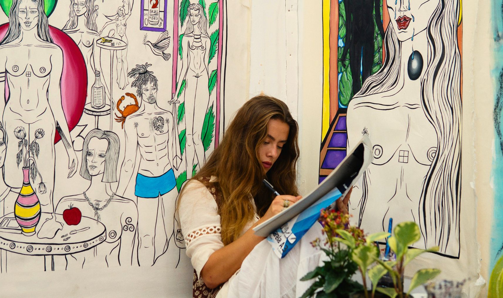 Kim Mac drawing on a sketchpad in front of a large painted mural of figures.